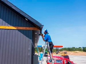Technician installing gutter guard mesh on metal building gutter from elevated ladder