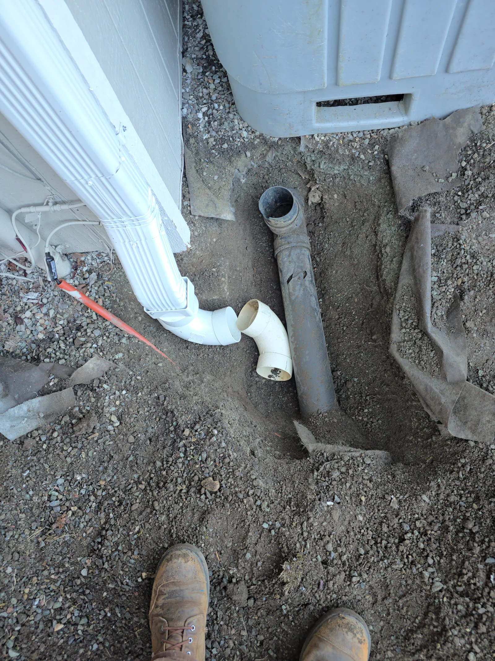 White vinyl downspout with elbow connector and metal drainage pipes on gravel foundation